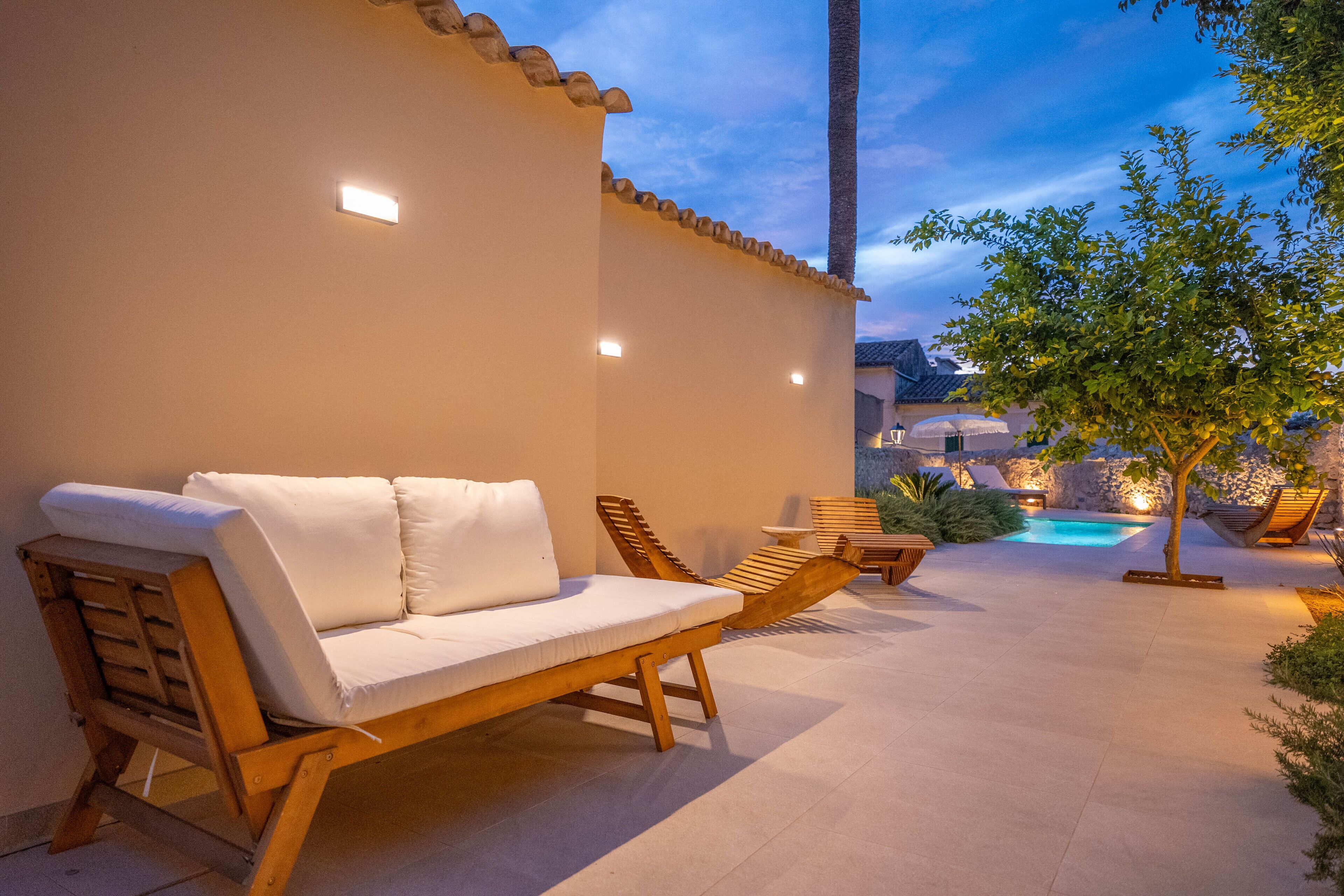 A patio with a wooden lounge chair and a sofa, a small table, and a tree.