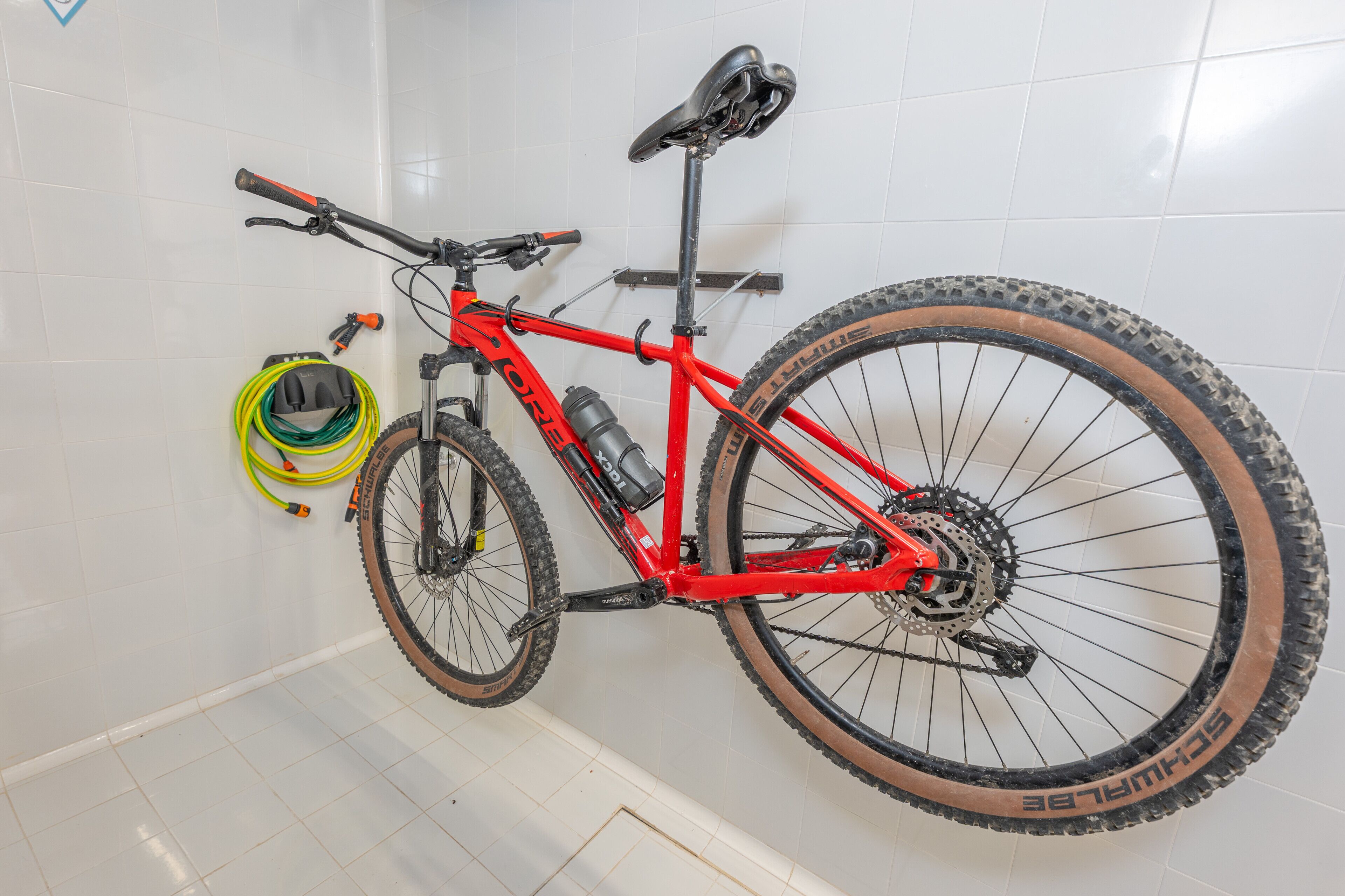 Una bicicleta de muntanya vermella amb una ampolla d'aigua i un casc, recolzada contra una paret de rajola blanca.