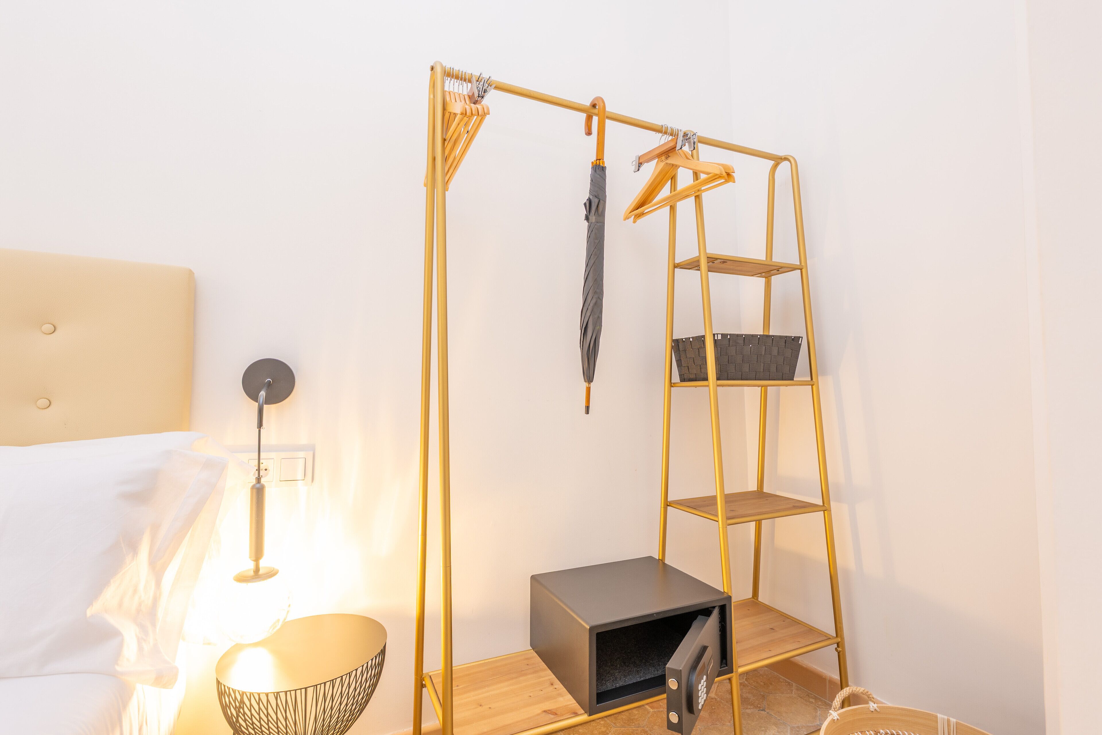 A room with a bed, a bedside lamp, a small table with a bowl, a wall-mounted shelf, and a hanging rack.