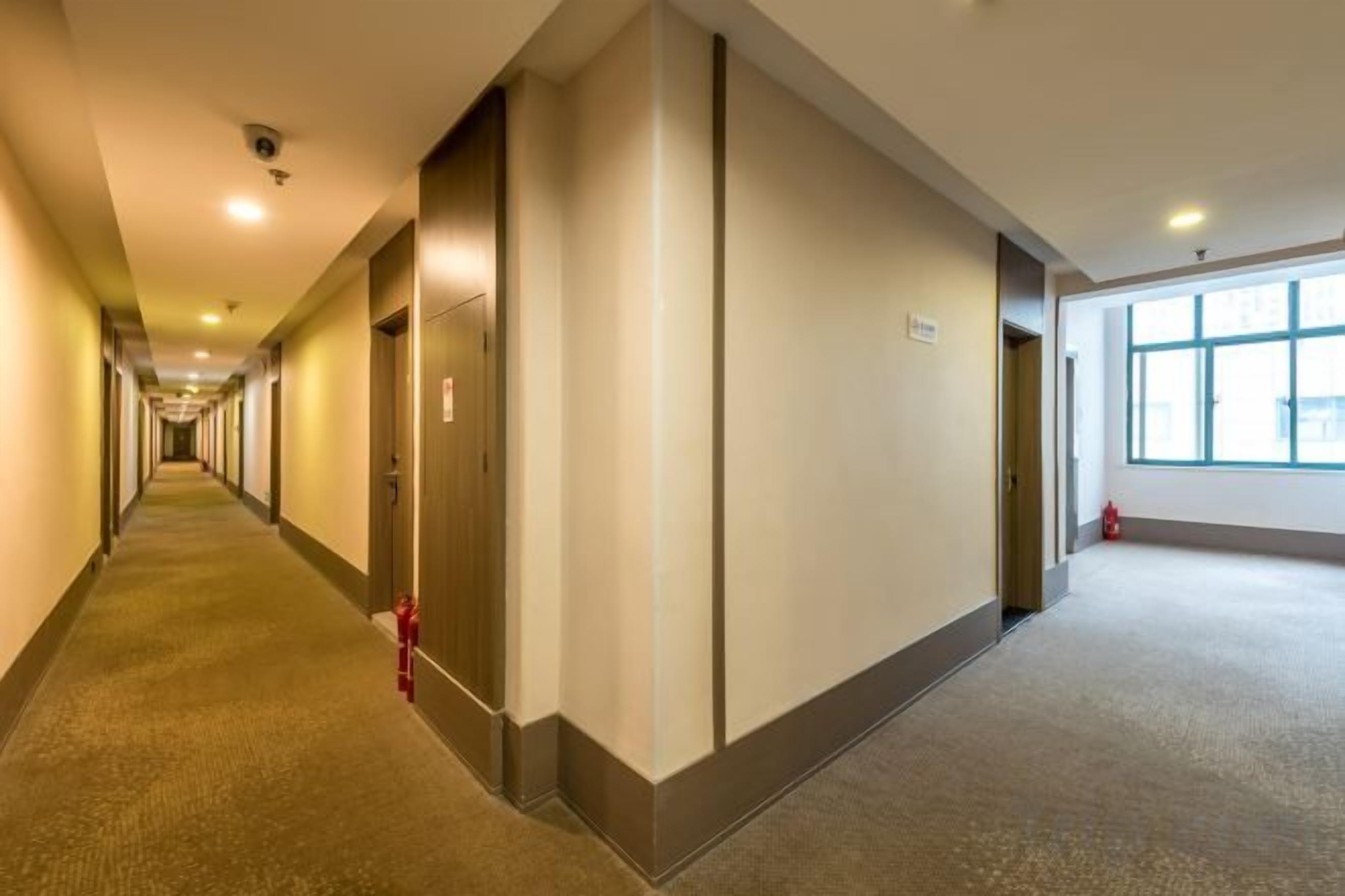 A long, narrow hotel corridor with doors on both sides, a fire extinguisher on the left, and a window on the right.
