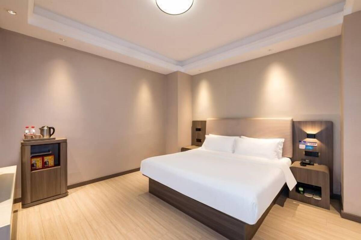 Habitación de hotel con una cama grande, mesitas de noche, una lámpara de pared y un mueble pequeño con una tetera.
