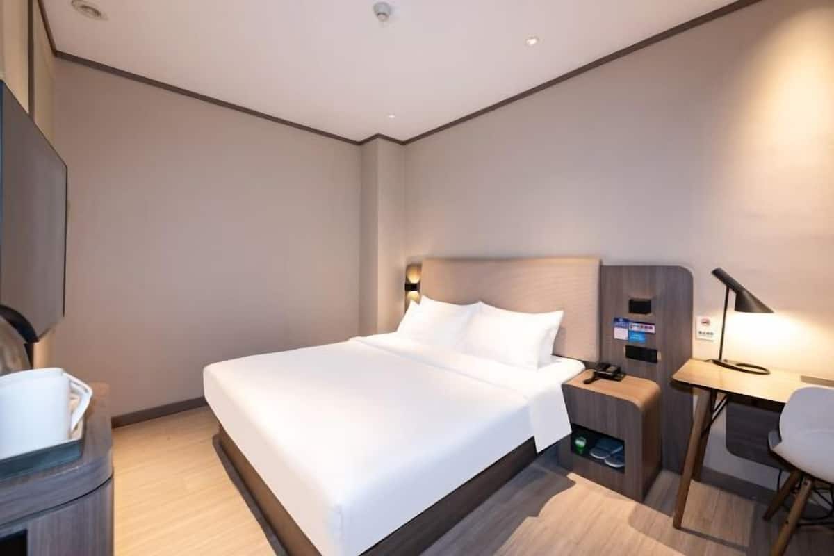 Habitación de hotel con una cama grande, una mesita de noche con lámpara, un escritorio pequeño y una silla.