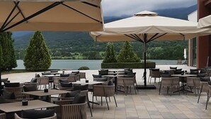 Un patio al aire libre con mesas y sillas, sombrillas grandes y vistas a un lago y montañas.