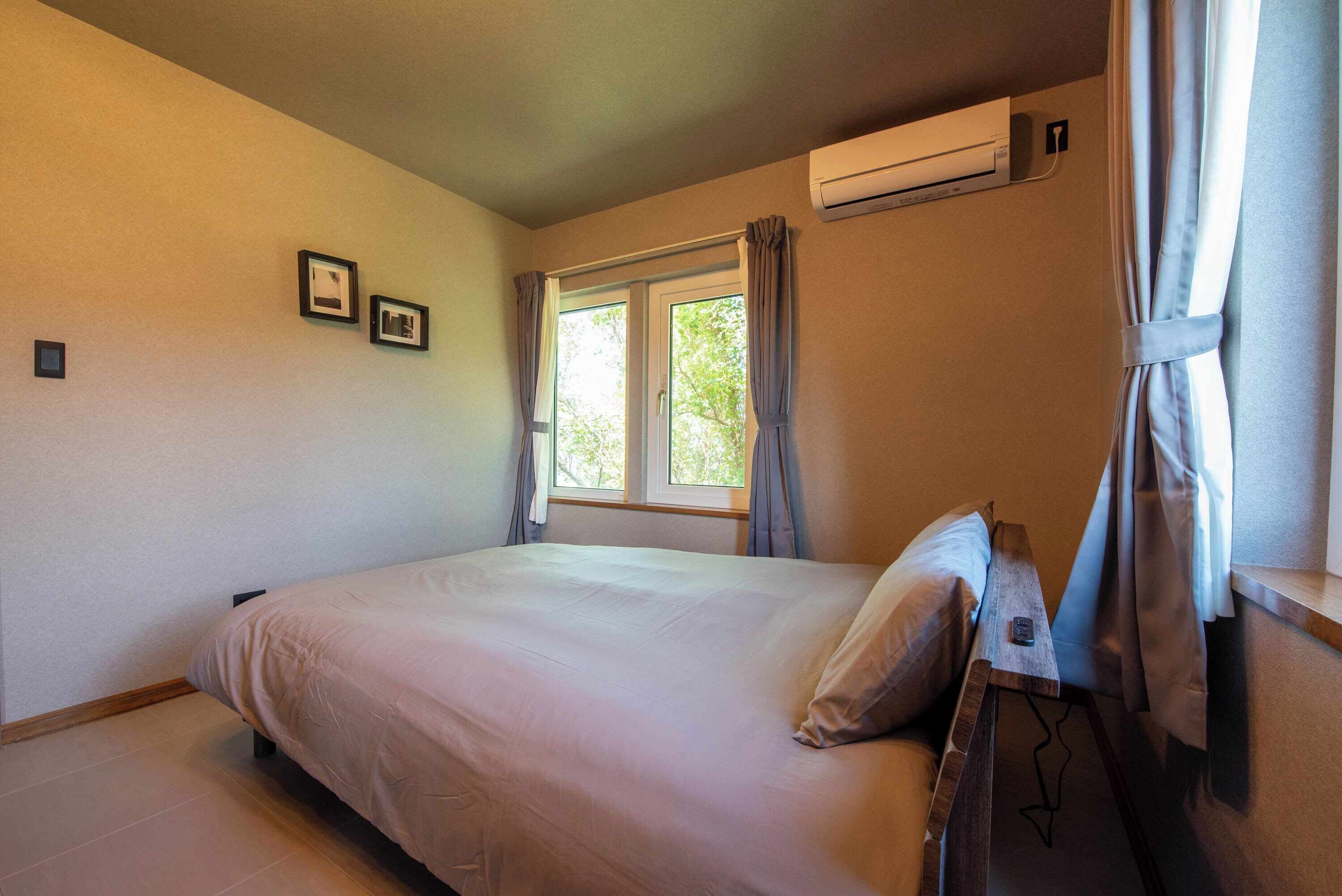 A bedroom with a bed, a window with curtains, an air conditioning unit, and a small table with a lamp.