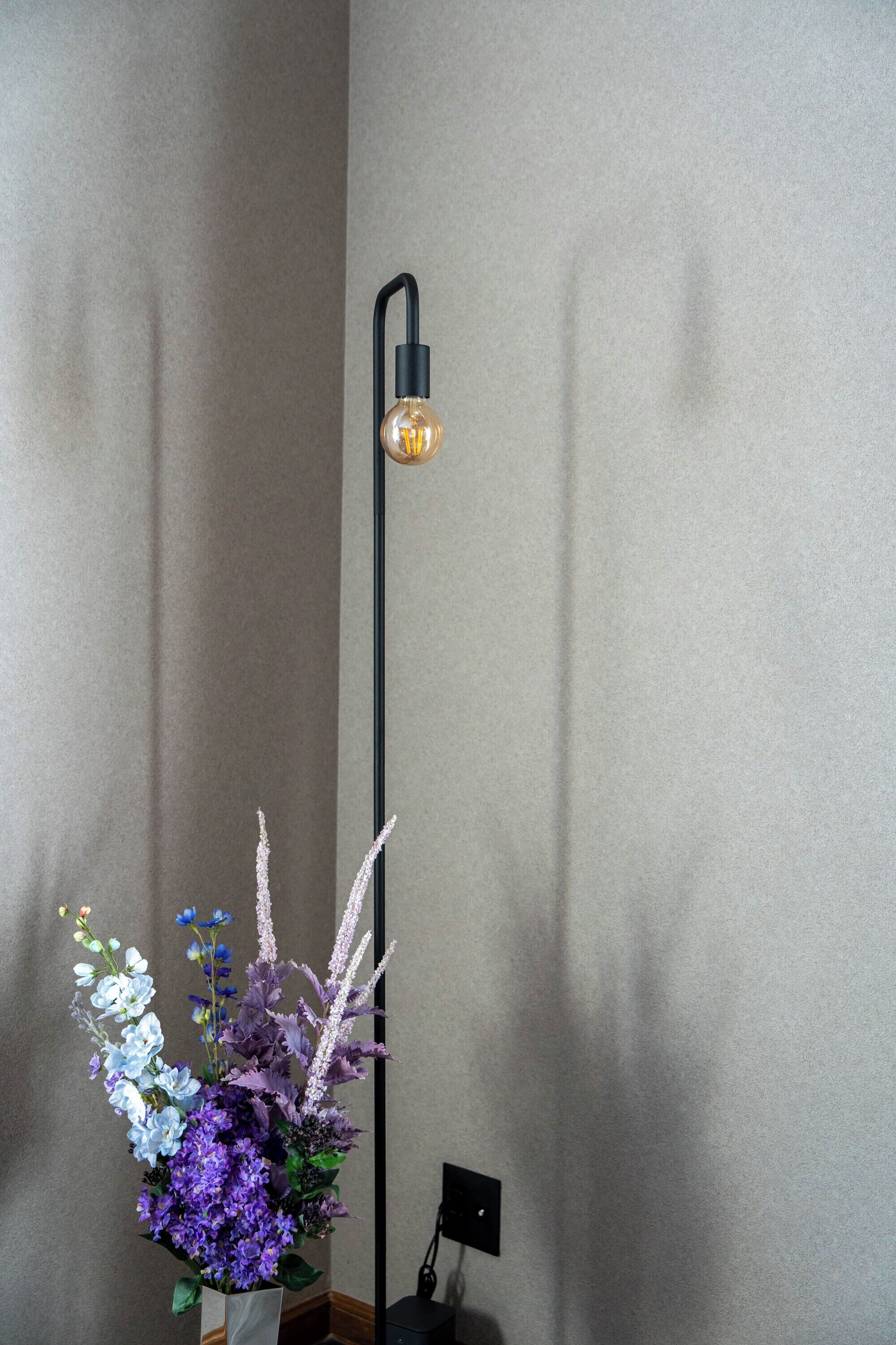 A tall floor lamp with a black base and a light bulb, positioned next to a vase of flowers with various colors.