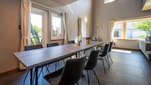 Ein Essbereich mit einem langen Tisch, Stühlen und Vorhängen. Es gibt Fenster mit Vorhängen, ein Sofa und eine Lampe auf dem Tisch.