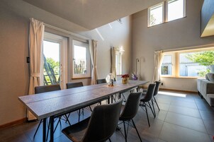 A dining area with a long table, chairs, and curtains. There are windows with curtains, a sofa, and a lamp on the table.