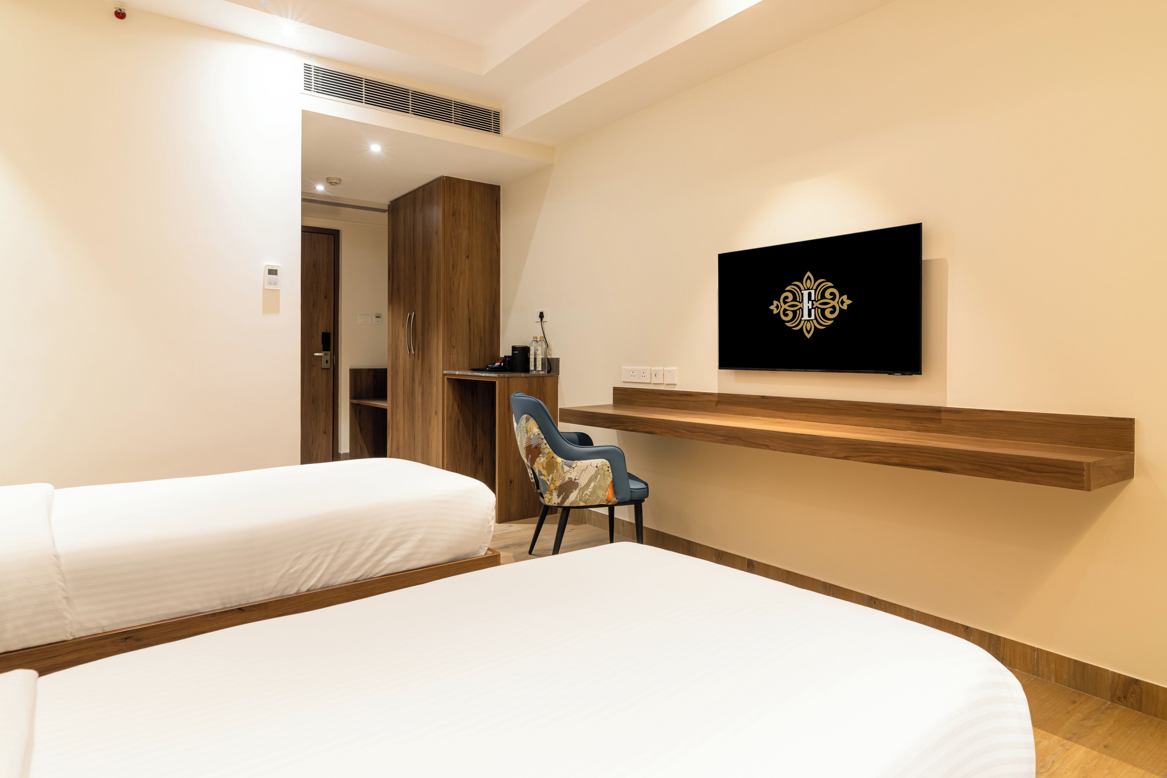 A hotel room with two beds, a desk, a chair, and a wall-mounted TV.