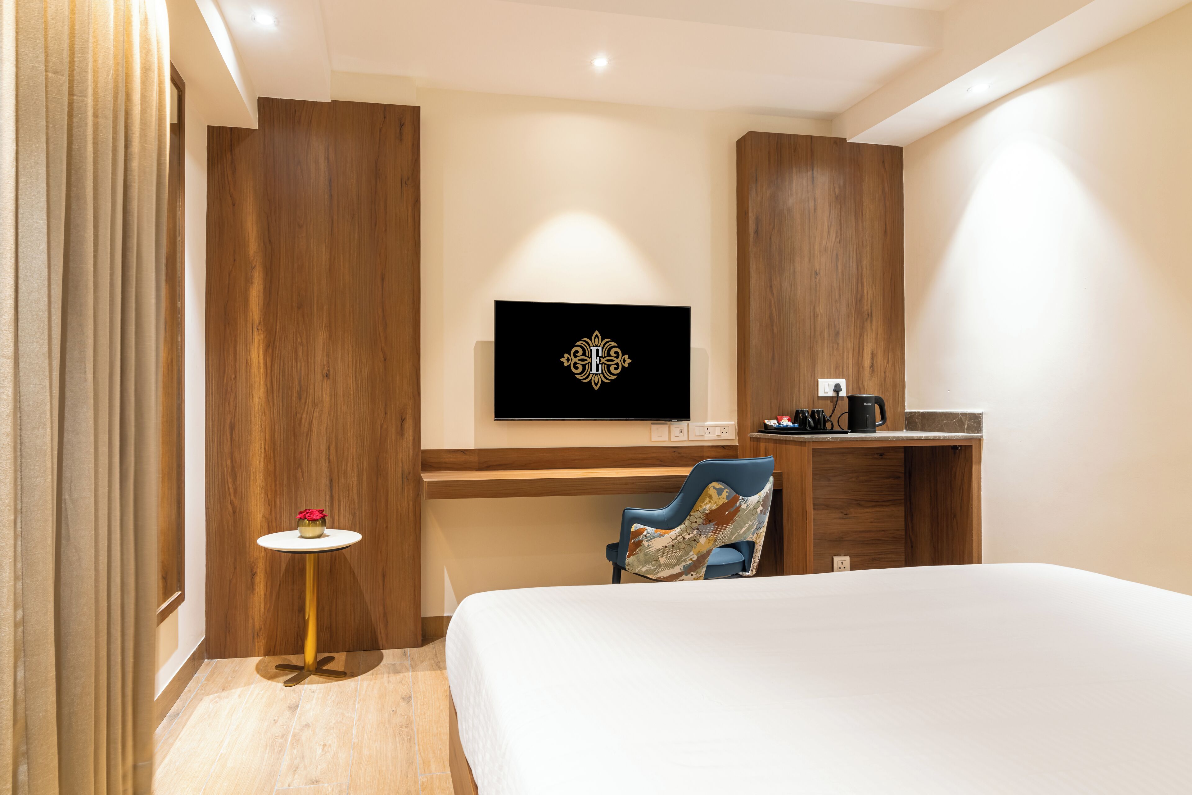 A hotel room with a bed, a desk, a chair, a small table with a flower, and a TV on the wall.
