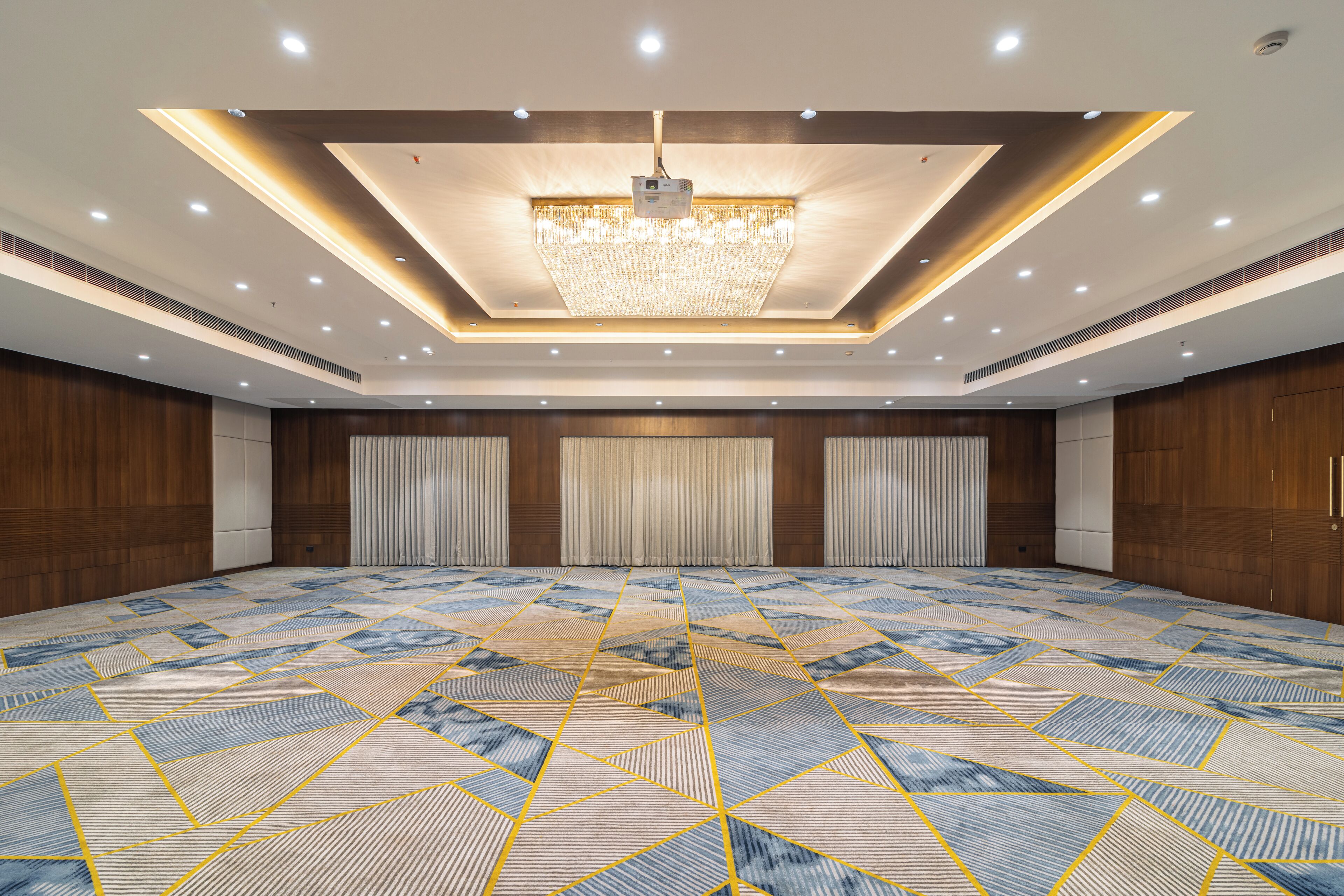 Een ruime congreszaal met een geometrisch patroon in het vloerkleed, in het plafond weggewerkte verlichting en een centrale kroonluchter.