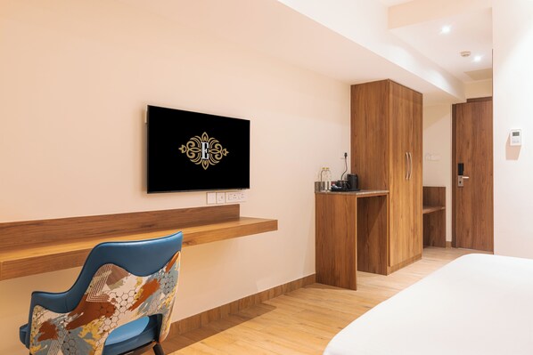 A modern hotel room with a wooden wardrobe, a flat-screen TV mounted on the wall, a blue chair with a patterned cushion, and a wooden bench. - Hotel Royal Embassy (Erode)