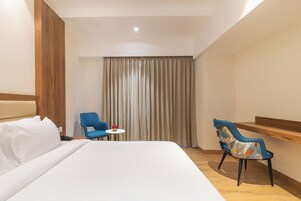 Una habitación de hotel con una cama grande, dos sillas, una mesita con un florero y un estante de madera.