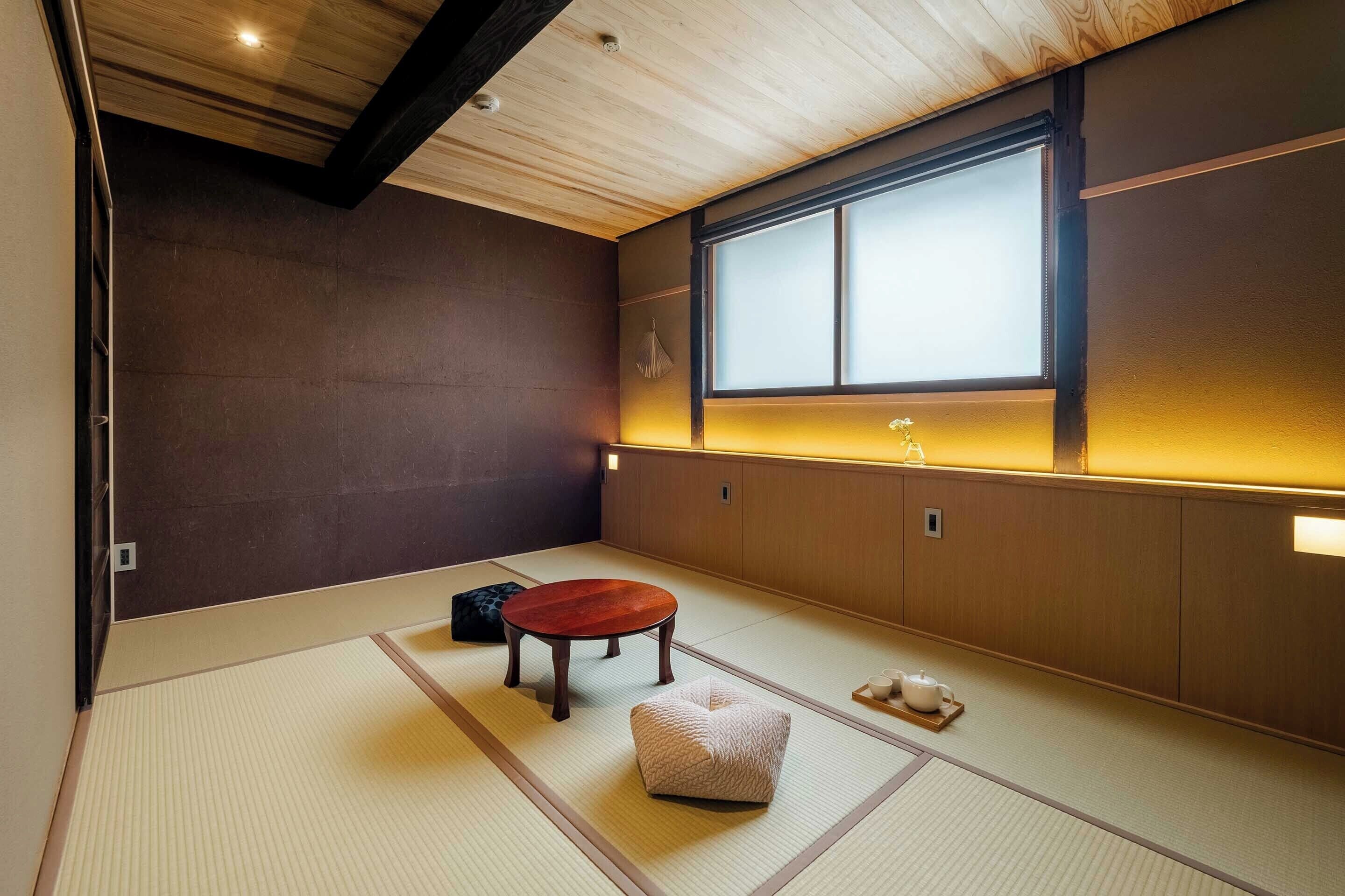 A tatami mat room with a wooden ceiling, a sliding door, a tatami mat table, a small tatami mat stool, and a window with a view.