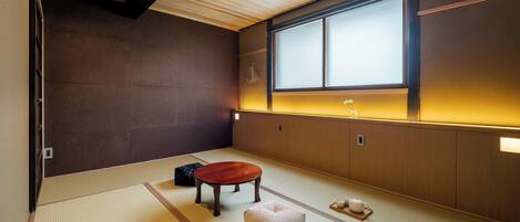 A tatami mat room with a wooden ceiling, a sliding door, a tatami mat table, a small tatami mat stool, and a window with a view.
