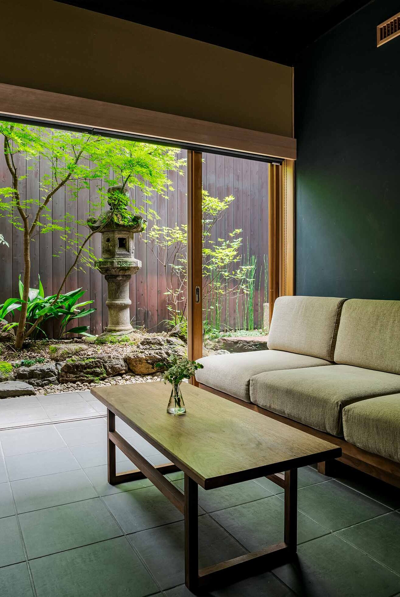 A modern living room with a sofa, a wooden coffee table, and a view of a garden with a stone lantern and bamboo plants.