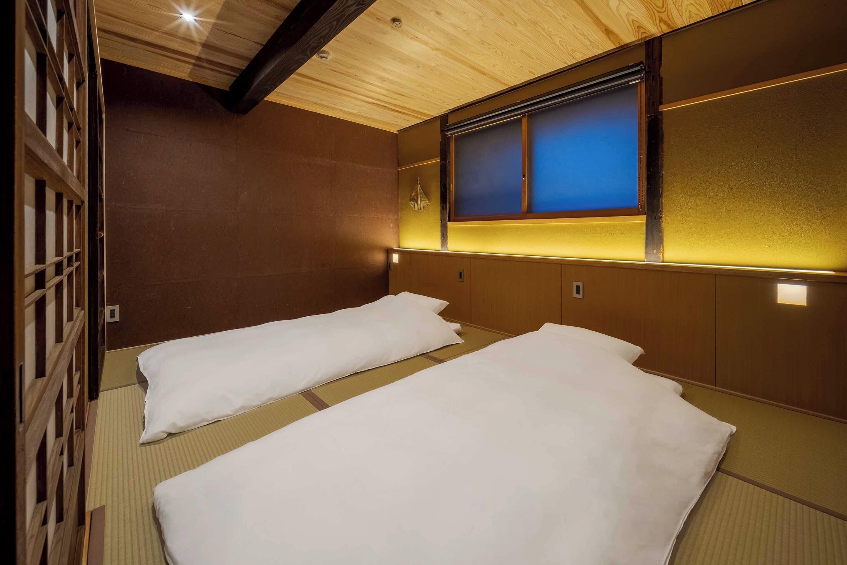 Une chambre avec deux lits, des murs en bois, une fenêtre teintée de bleu et un sol recouvert de tatamis, ces nattes traditionnelles japonaises.