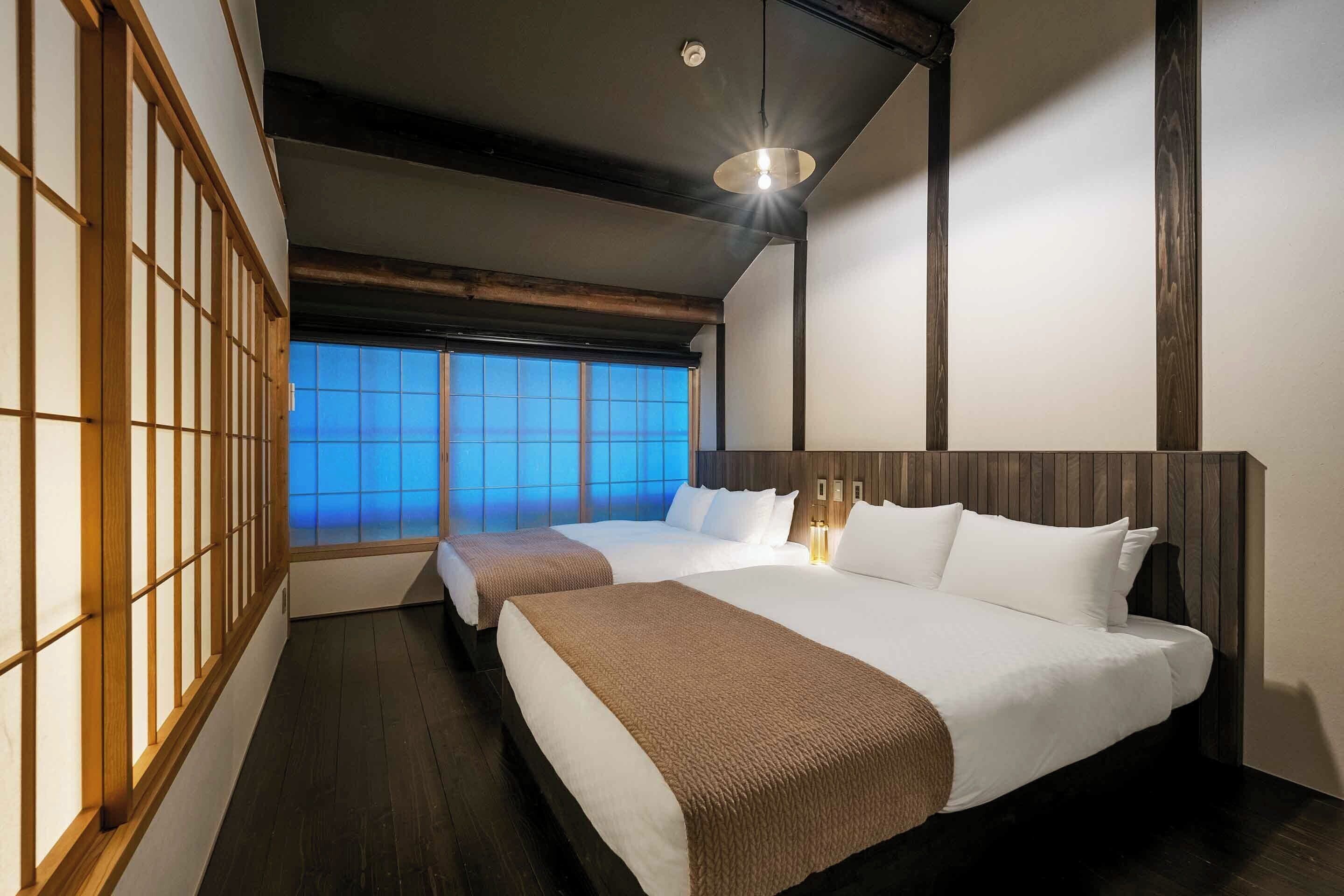 A hotel room with two beds, wooden flooring, a large window with blue sky view, and a modern ceiling light.