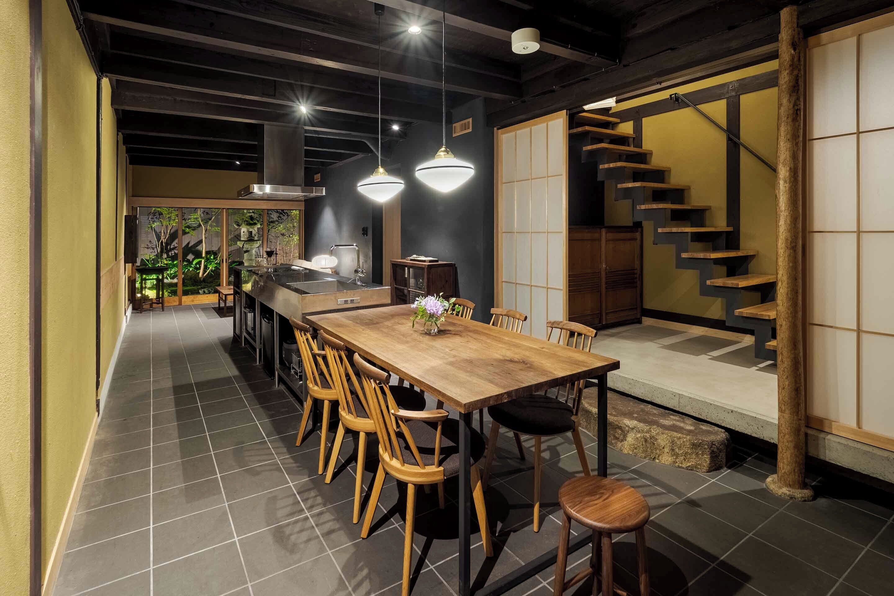 A spacious dining area with a long wooden table and chairs, a kitchen in the background, and a traditional staircase.
