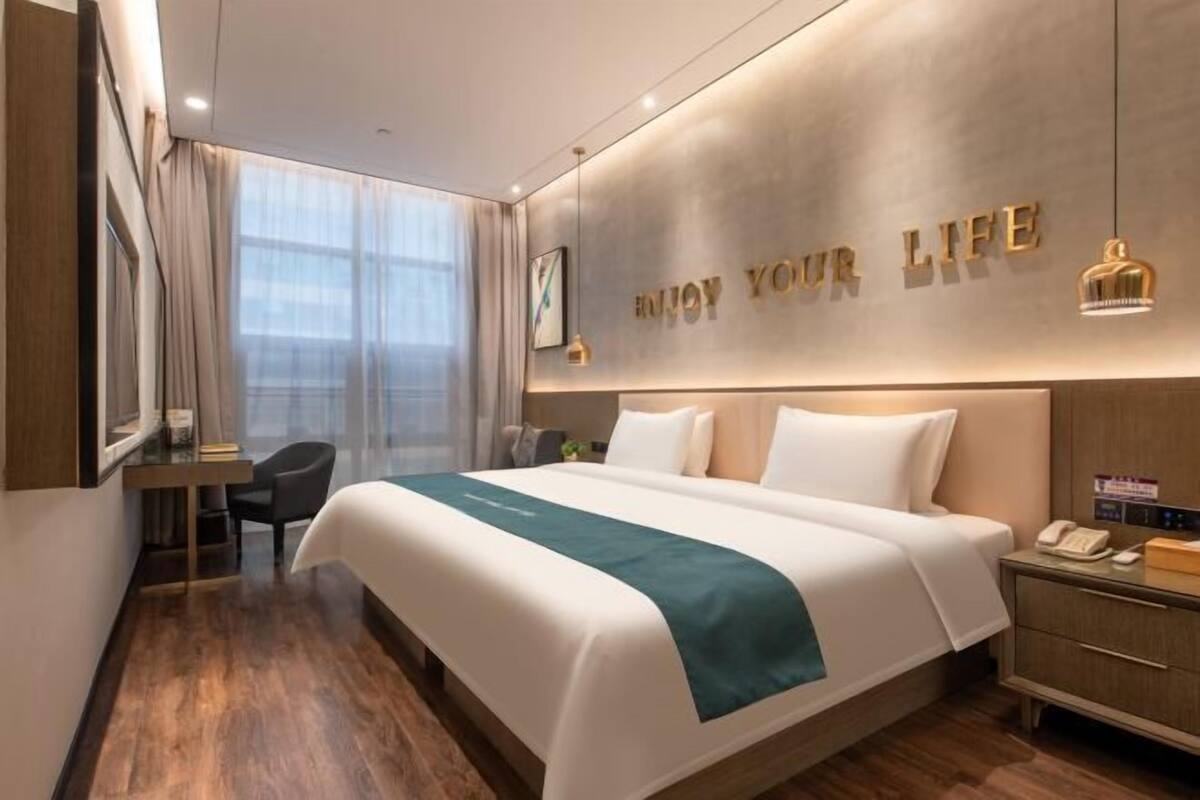 A hotel room with a large bed, a desk, a chair, a nightstand, and a wall with the text 'ENJOY YOUR LIFE'.