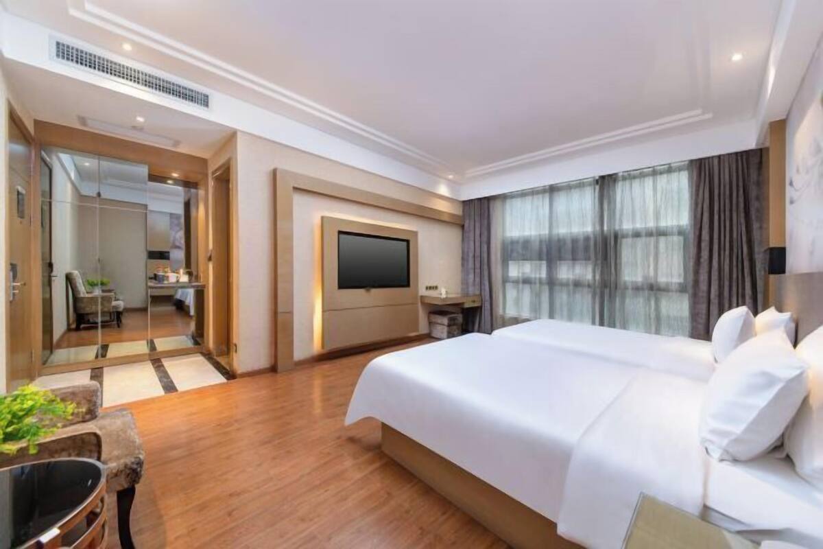 A hotel room with a large bed, a flat-screen TV mounted on the wall, a desk with a chair, and a view of the city through large windows.