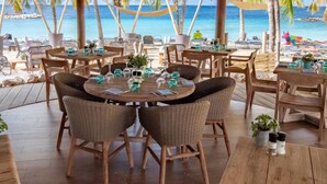 En restaurang vid stranden med runda bord, flätade stolar och krukväxter.