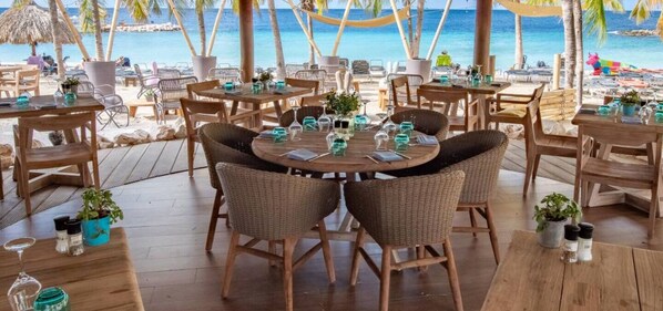 A beachside restaurant with round tables, wicker chairs, and potted plants. - VIP Blue Bay Beach Villa 34 (Sint Michiel)