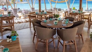A beachside restaurant with round tables, wicker chairs, and potted plants. - VIP Blue Bay Beach Villa 5 (Sint Michiel)