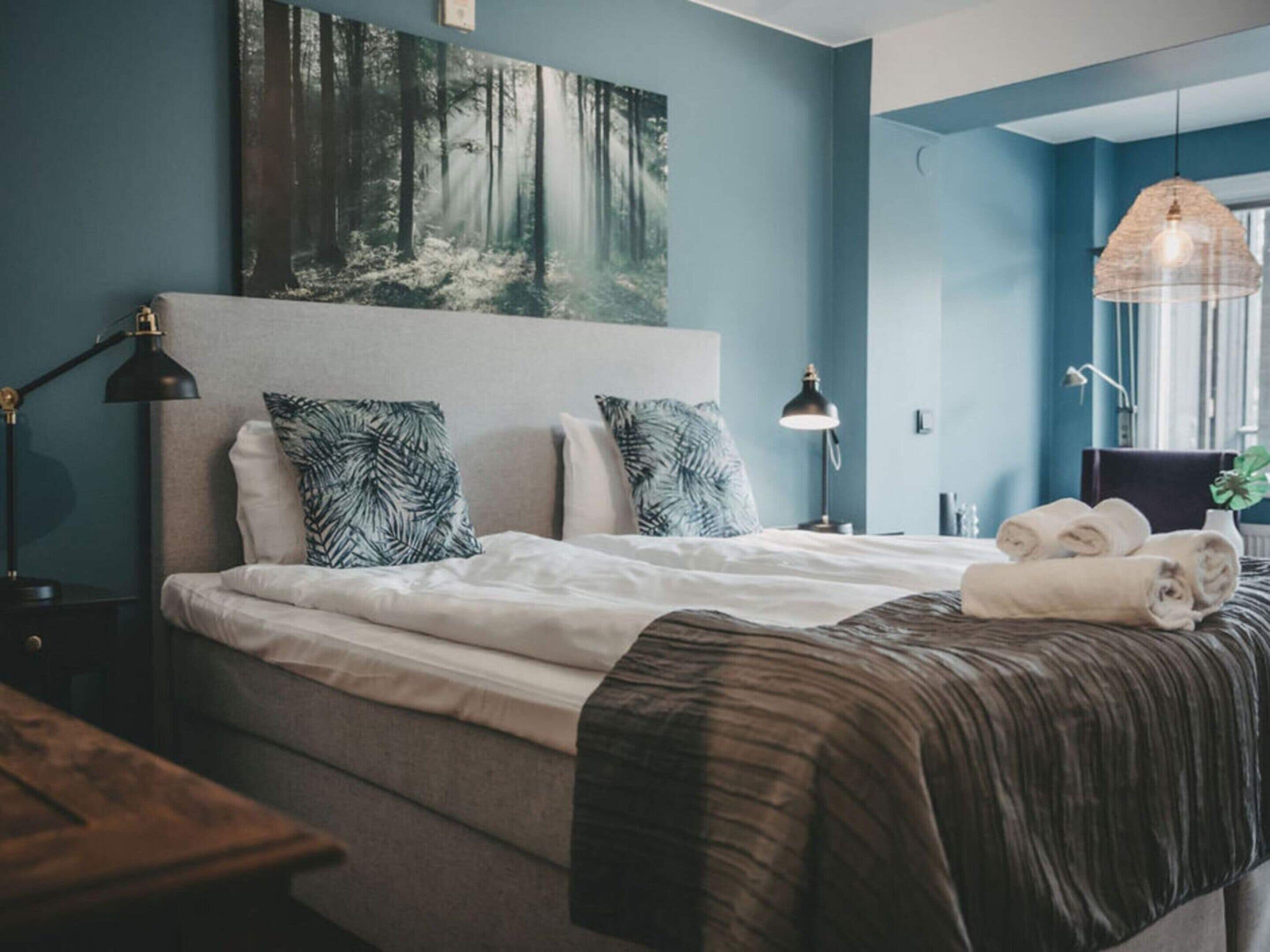 A bedroom with a bed, pillows, a headboard, bedside lamps, a hanging light fixture, and a painting on the wall.