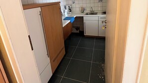 Eine schmale Küche mit weißen Schränken, einem Kühlschrank und einem Fliesenboden.