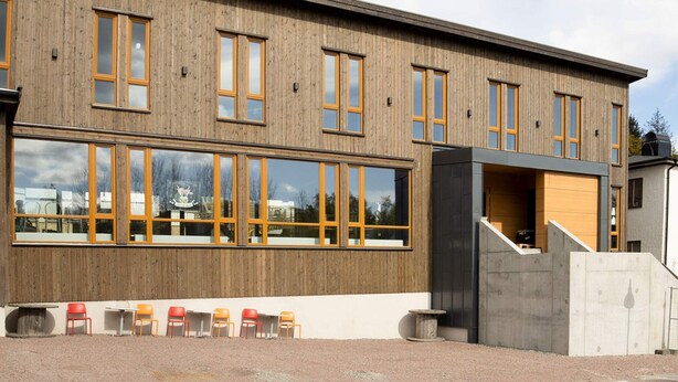 En moderne bygning med facade i træ, store vinduer og et overdækket udendørs siddeområde.