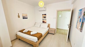 A bedroom with a wooden bed, white linens, a small bedside table, and a door leading to another room. - Seaside Comfort Residence (Larnaca)