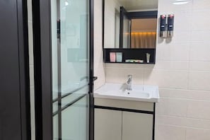 Un baño moderno con mampara de vidrio, un lavabo blanco, un espejo y un estante con artículos de aseo.