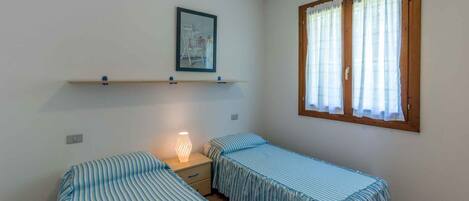 Una habitación pequeña y limpia con dos camas individuales, una mesita de noche, un estante con un cuadro enmarcado y una ventana con cortinas.
