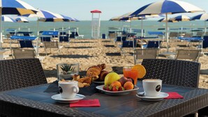 A beachside breakfast table with croissants, fruit, and coffee, overlooking the beach with umbrellas and lounge chairs. - Houses and Apts. in Albarella 24882 (Rosolina)