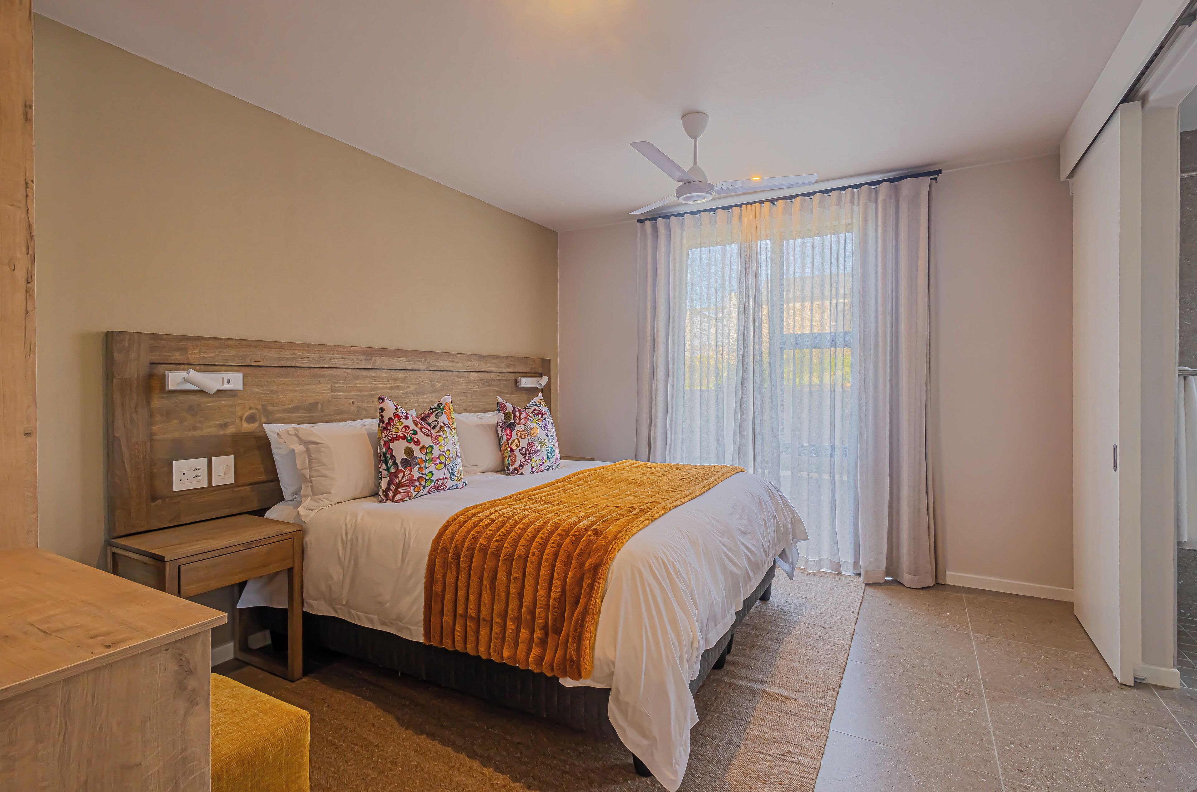 A bedroom with a wooden headboard, a bed with white bedding and a brown throw, a nightstand, a bedside table, and a window with sheer curtains.