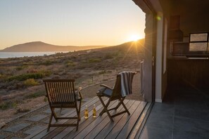 Terraza de madera con dos sillas y dos botellas de cerveza, con vista a una playa y una puesta de sol, con montañas al fondo.