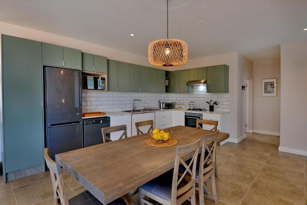 Una cocina moderna con armarios verdes, un refrigerador de acero inoxidable, una mesa de comedor de madera con sillas y una lámpara colgante.