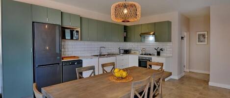 Una cocina moderna con armarios verdes, un refrigerador de acero inoxidable, una mesa de comedor de madera con sillas y una lámpara colgante.