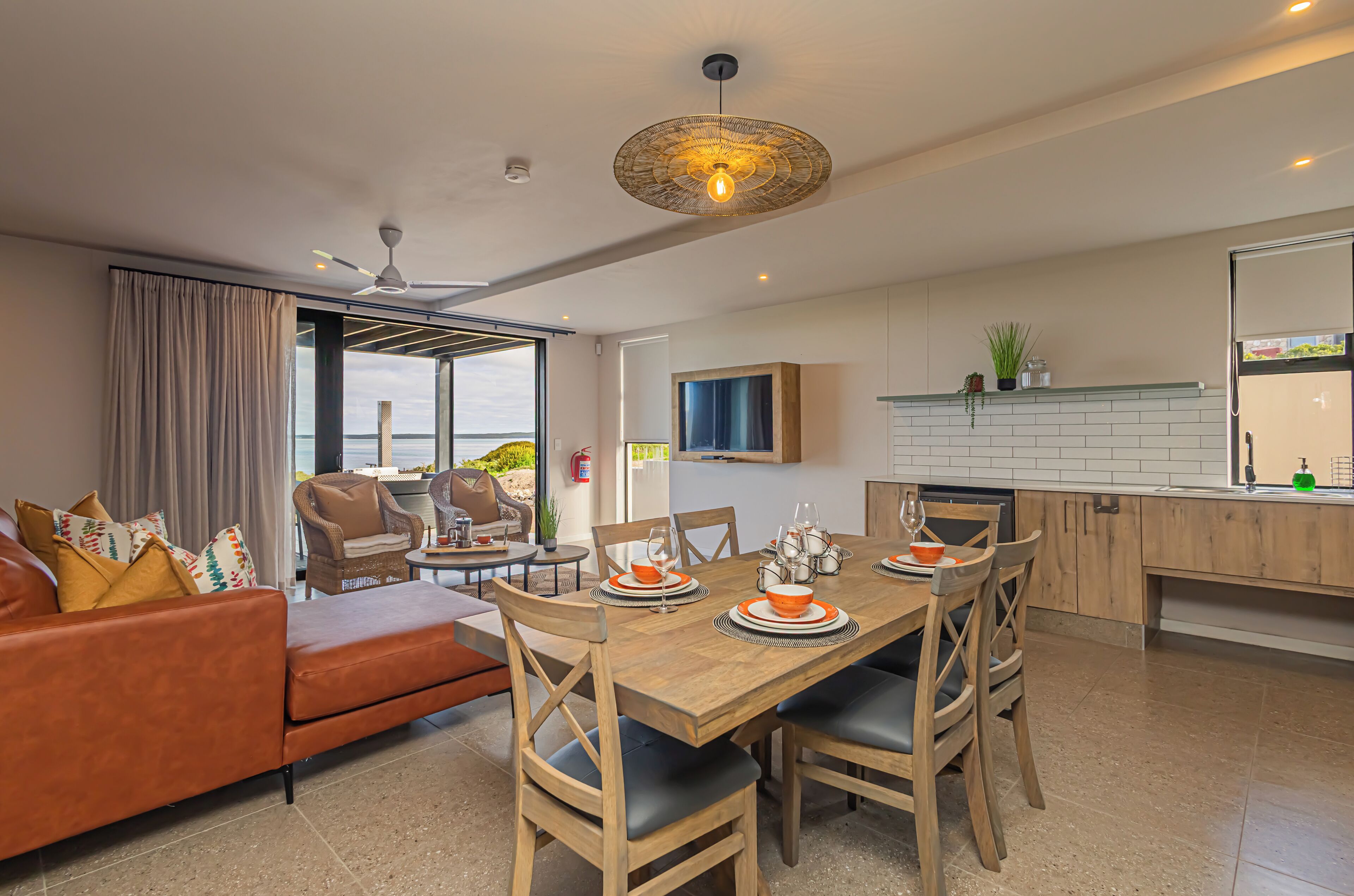 A modern living room with a wooden dining table set for four, a sofa with patterned cushions, and a ceiling fan.