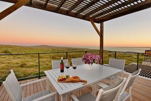 Mesa blanca dispuesta con copas de vino, una botella y una bandeja de frutas en una terraza con vista al mar.
