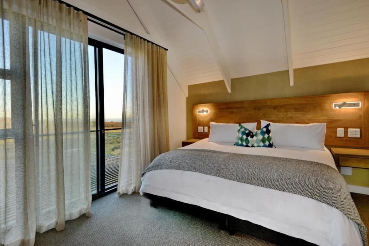 Habitación de hotel con una cama grande, cabecera de madera, mesita de noche y vistas a través de un ventanal.