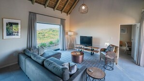 A modern living room with a sofa, coffee table, and TV, a dining area with chairs, and a large window offering a view of the outdoors. - The Shark Bay Private Villas (Langebaan)