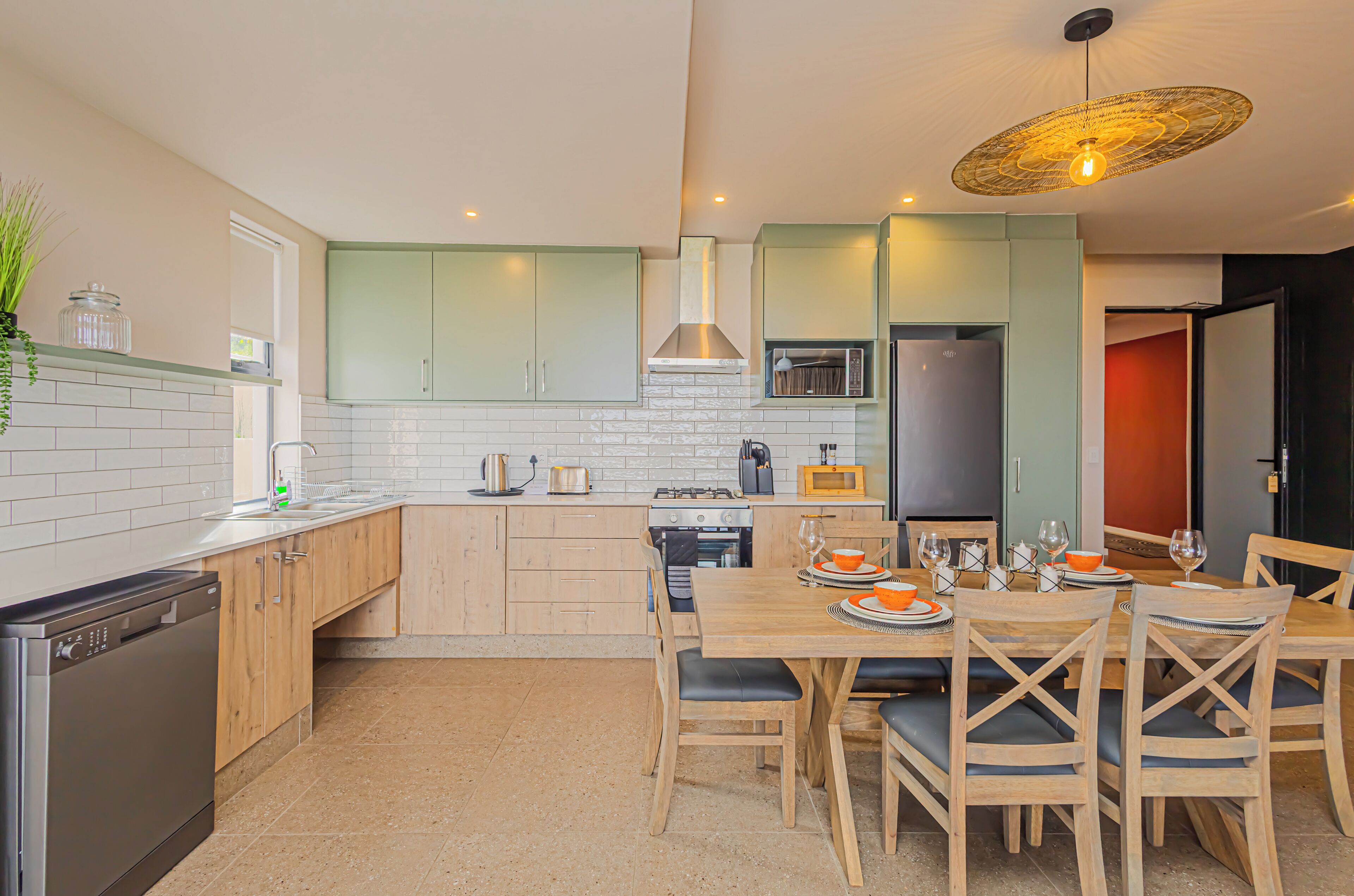 A modern kitchen with light green cabinets, a wooden dining table, and a hanging light fixture.