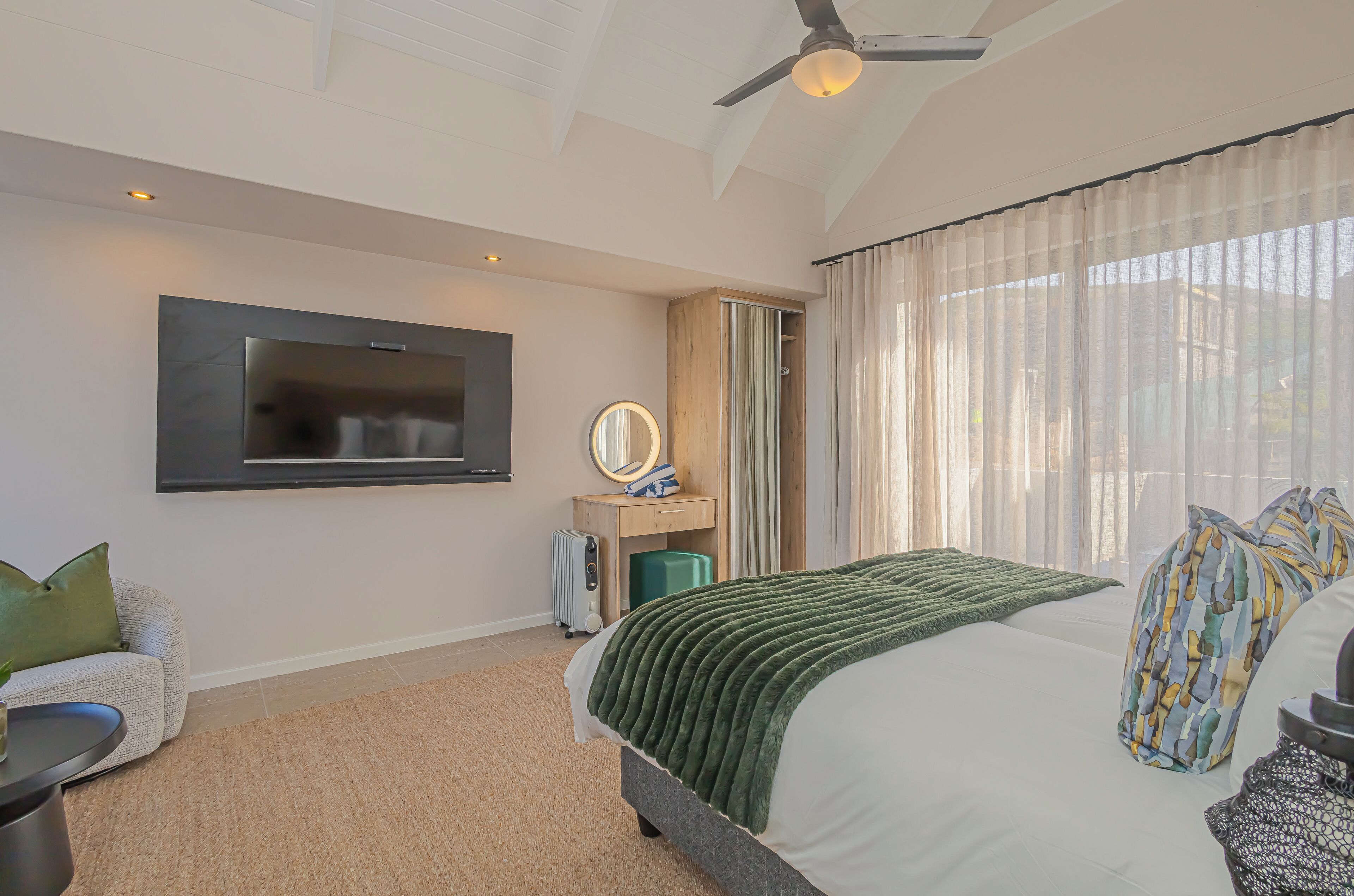 Une chambre moderne avec un grand lit, un téléviseur à écran plat fix é au mur, une table de chevet avec une lampe et une fenêtre ornée de rideaux légers.