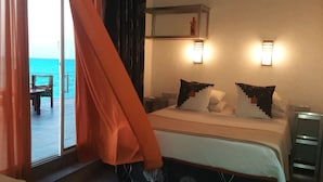A hotel room with a bed, two bedside tables, a nightstand, a window with curtains, a view of the sea, and a small shelf with a decorative item. - Game Fish Apartment (Nungwi)