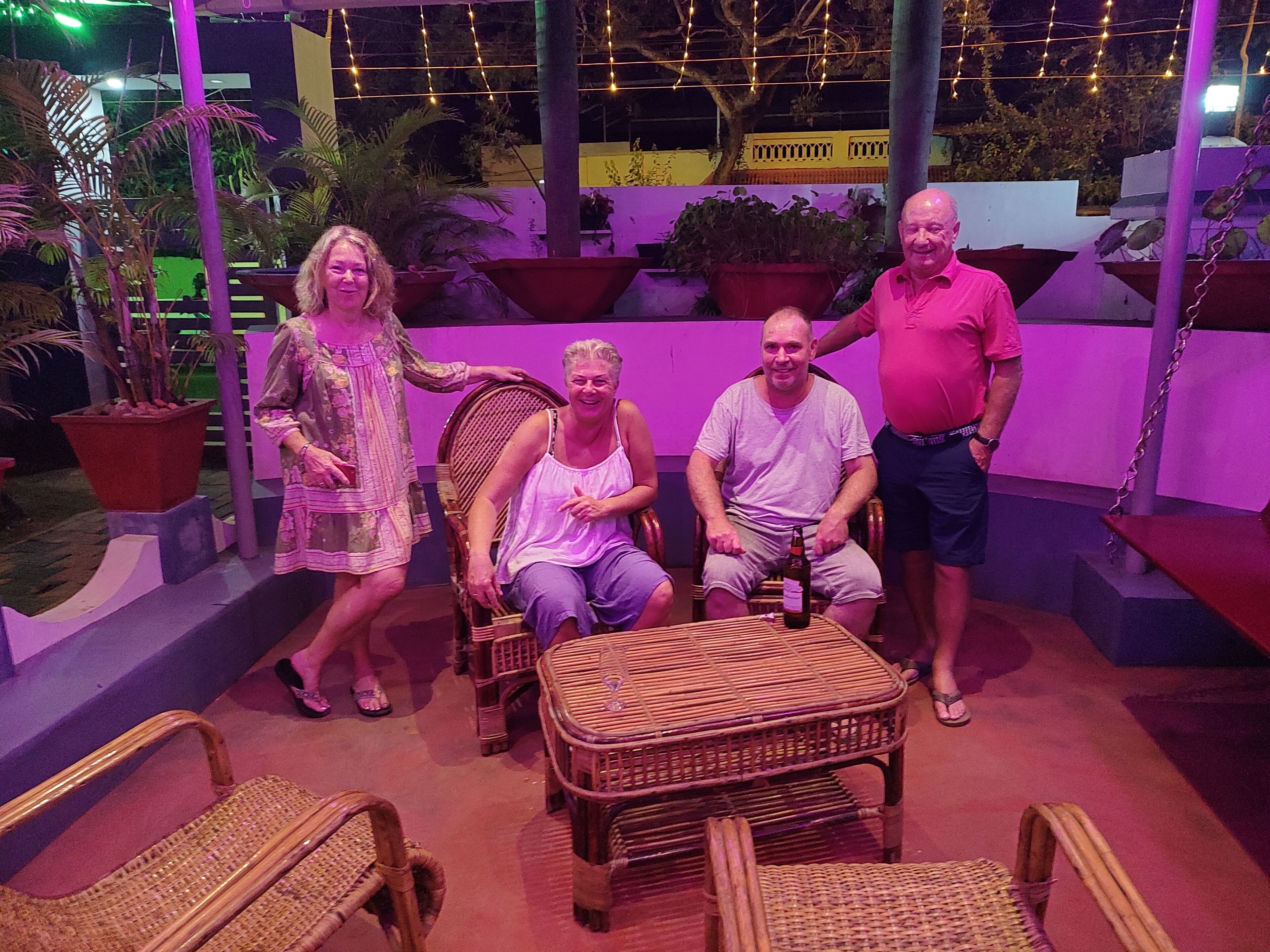 Un groupe de personnes est assis sur des chaises en osier autour d’une table, dans un espace extérieur agrémenté de plantes en pot et illuminé de guirlandes lumineuses.