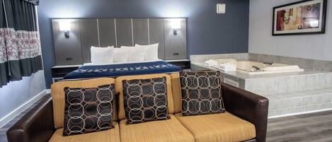 A hotel room with a bed, a sofa with patterned cushions, a large window with curtains, a bathroom with a bathtub, and a framed picture on the wall.