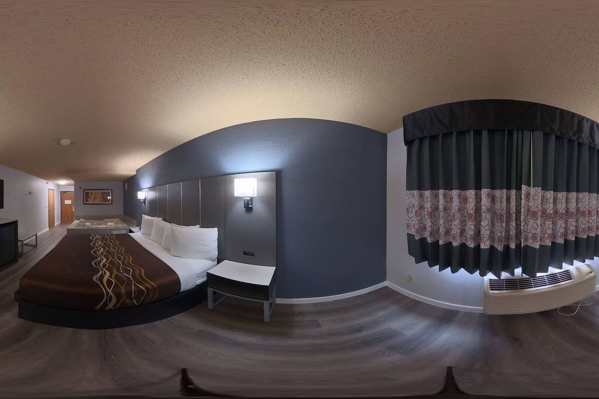 Une chambre d’hôtel moderne avec un plafond incurvé, un lit recouvert d’une couvre-lit à motifs, une table de chevet, un téléviseur fixé au mur et une fenêtre avec des rideaux.