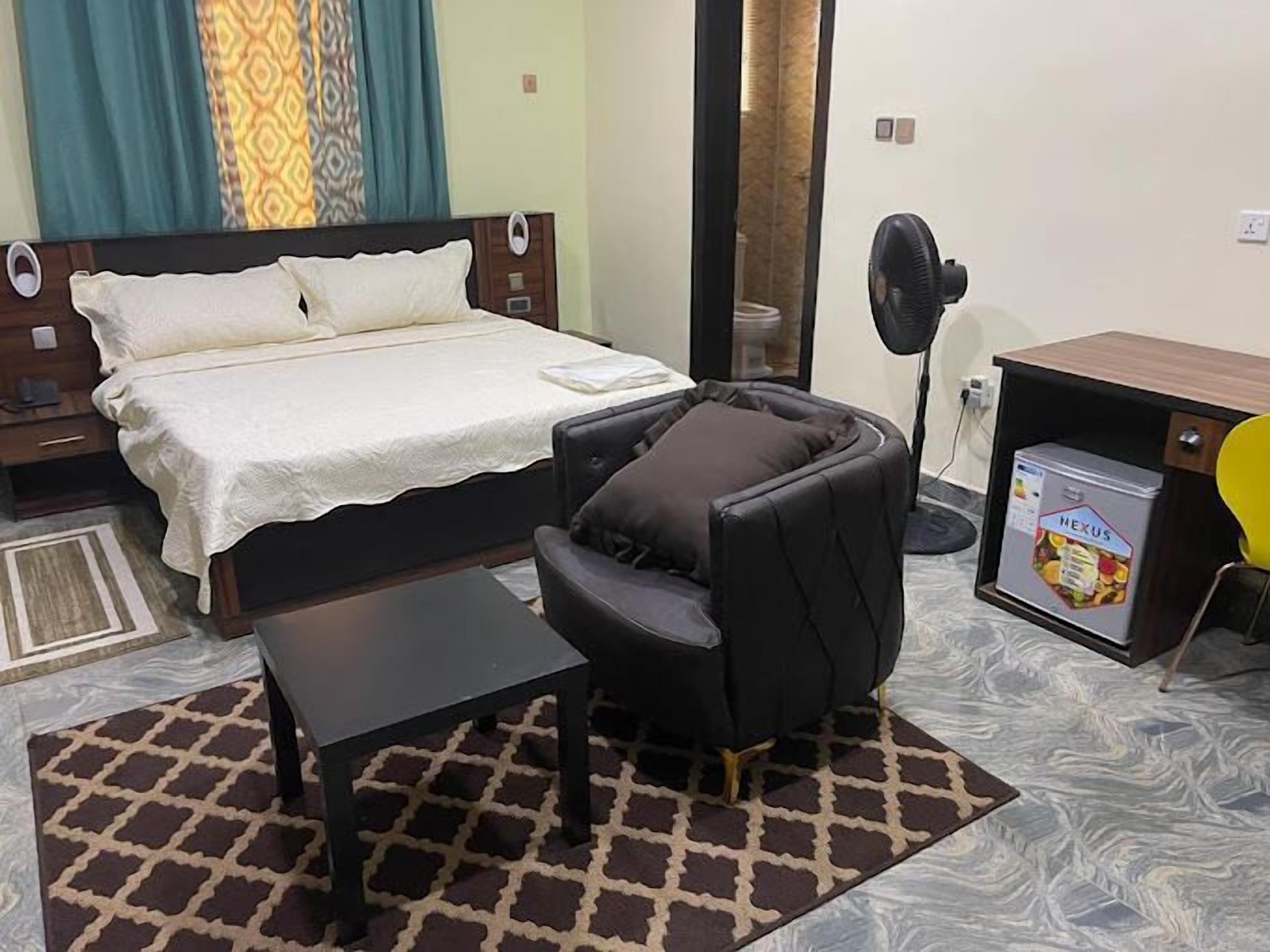 A bedroom with a bed, a small table, a massage chair, a desk with a fan, and a minibar.