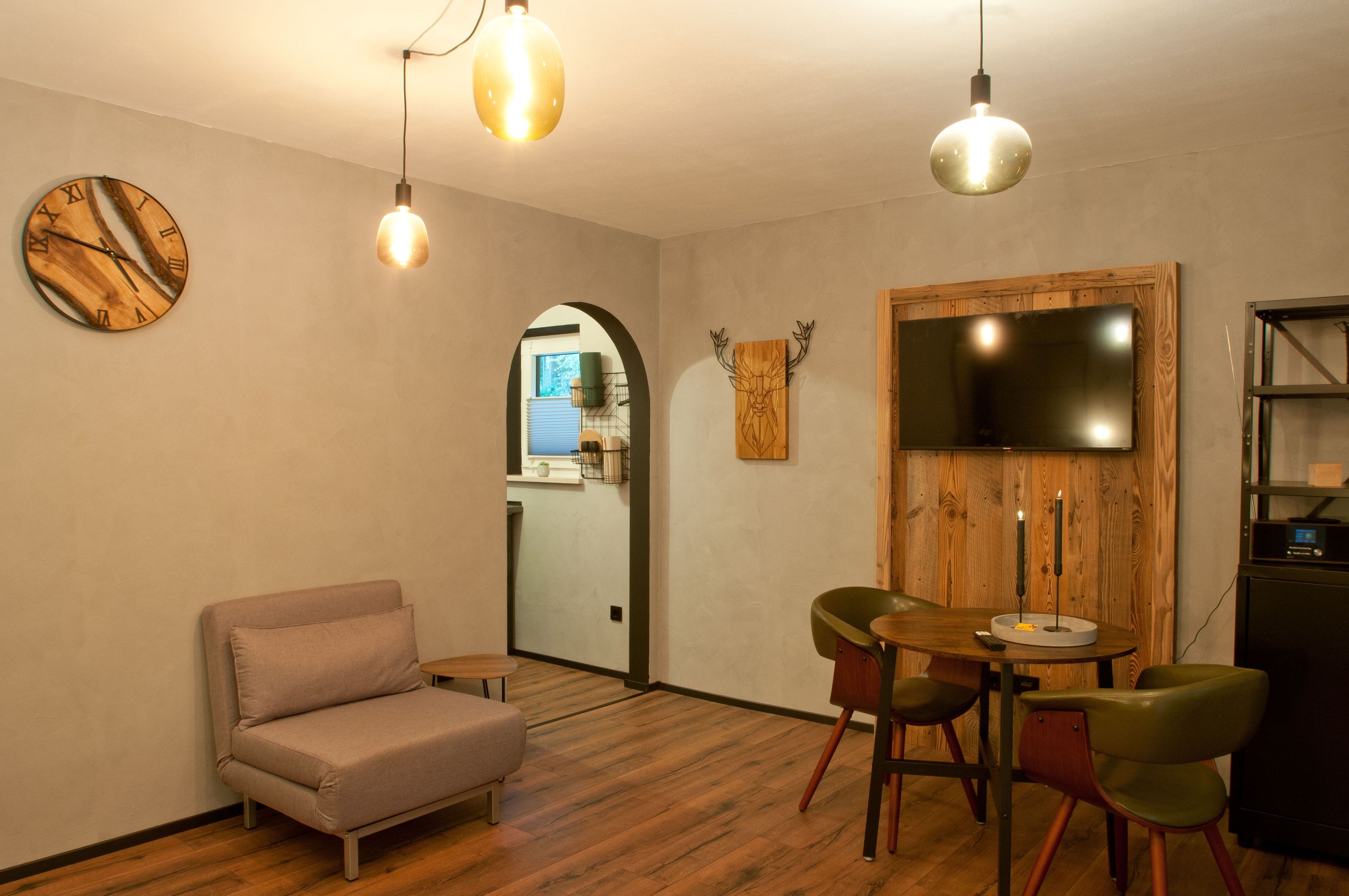 Een knus ingerichte kamer met een bank, een ronde tafel met stoelen, een houten wanddecoratie en een klok.